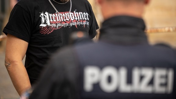 Ein Teilnehmer einer von einem NPD-Funktionär angemeldeten Demonstration trägt ein T-Shirt mit der Aufschrift "Ureinwohner" während er von einem Polizisten kontrolliert wird.