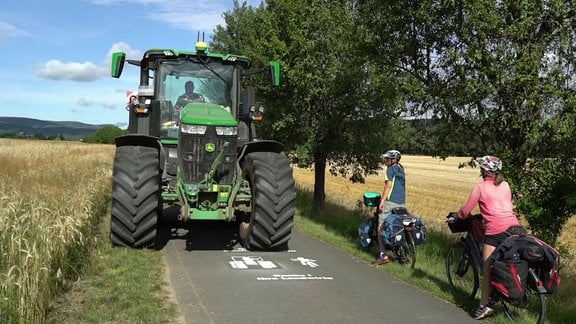 Zwei Fahrradfahrerinnen weichen einem Traktor auf einer Straße zwischen zwei Feldern aus.