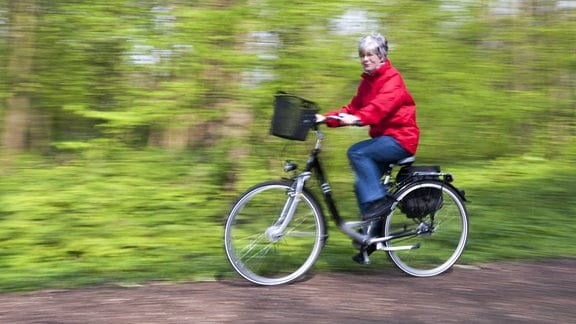 Seniorin in einer roten Jacke fährt Fahrrad durch den Wald