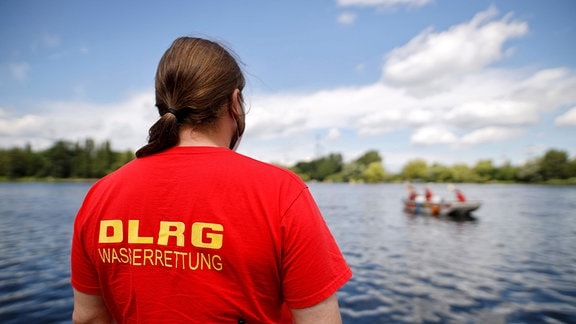 Eine Person mit einem T-Shirt der Deutschen Lebens-Rettungs-Gesellschaft (DLRG) steht mit dem Rücken zur Kamera an einem See.