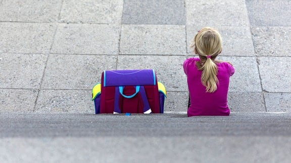Rückenansicht eines kleinen Mädchens mit Schulranzen auf einer Treppe sitzend.