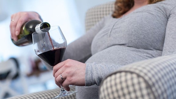 Eine schwangere Frau sitzt in einer Wohnung und giesst sich Rotwein in ein Glas (gestellte Szene).