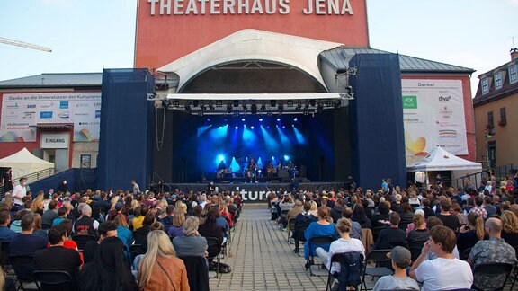 Blick aus dem Publikum auf eine blau beleuchtete Bühne, auf der Menschen musizieren