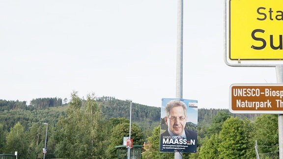 Ortseingangsschild von Suhl, im Hintergrund sind Wahlplakate und eine bergige Waldlanschaft zu sehen.