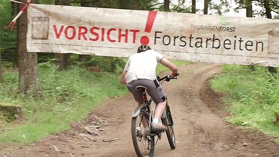 Ein Fahrradfahrer fährt auf einem Waldweg auf ein Banner zu auf dem "Vorsicht Forstarbeiten" steht. 