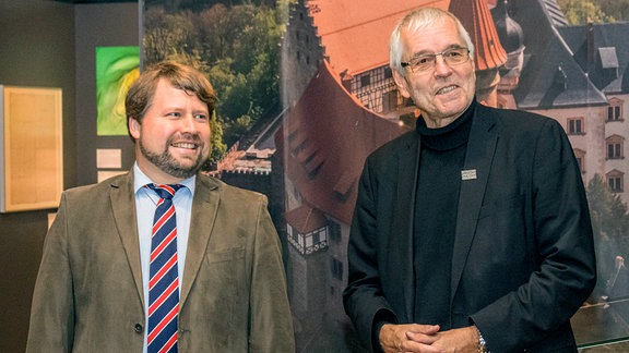 Ein Mann mittleren Alters steht neben einem älteren Mann mit weißem Haar und Brille.