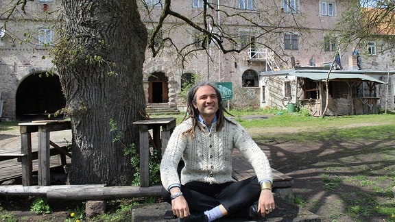Ein Mann sitzt lächelnd unter einem Baum auf dem Boden, im Hintergrund die hellgraue Fassade eines Schlosses