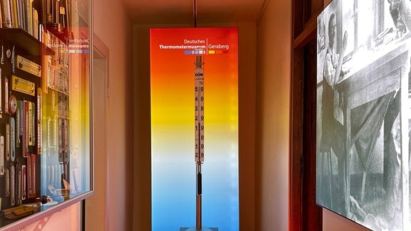 Ein menschengroßes Thermometer vor einem regenbogenfarbenen Hintergrund.