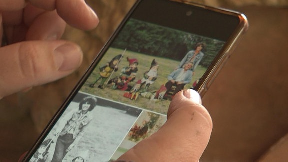 Eine Hand hält ein Handy. Auf dem sind alte Fotos von Menschen mit Gartenzwergen zu sehen.
