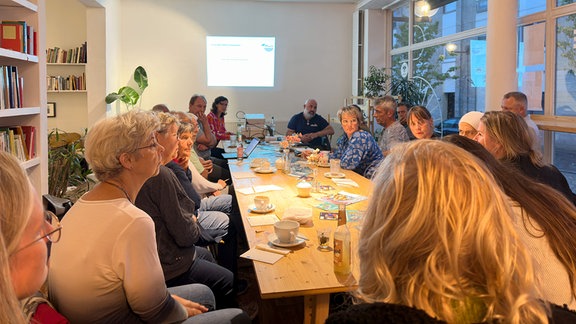 Mehrer menschen sitzten an einem Informationsabend um einen großen Tisch. im Hinterrund läuft eine Präsentation.