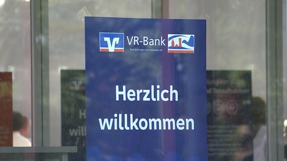 Ein Schild mit VR Bank Logos an einer Glastür auf dem "Herzlich Willkommen" steht.