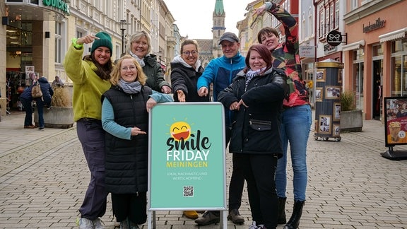 In einer Meininger Einkaufsstraße stehen Händlerinnen und Händler um einen Aufsteller, auf dem Smile Friday Meiningen zu lesen ist.