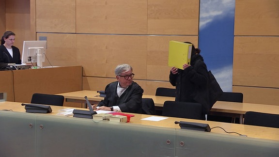 Eine Frau hat beim Betreten des Gerichtssaals ihr Gesicht hinter einem Ordner verborgen.  