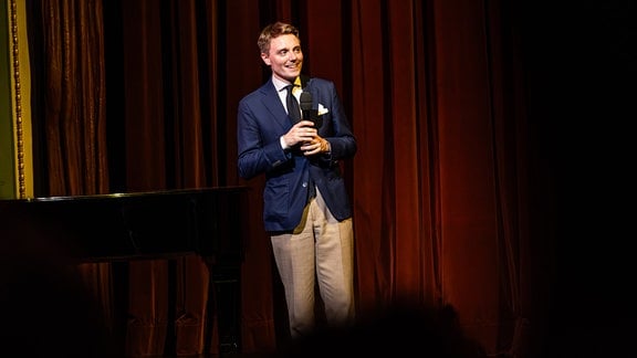 Ein Mann lächelnder Mann steht mit Mikrofon auf einer Bühne.