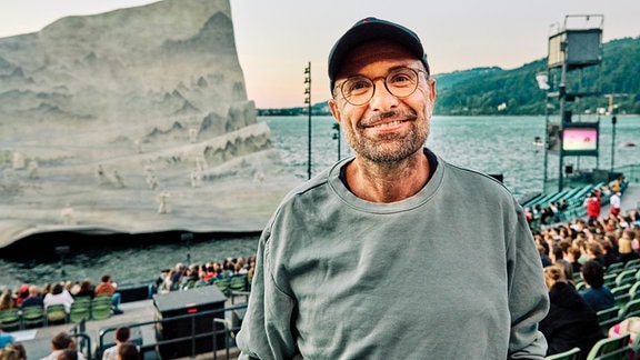 Regisseur Philipp Stölzl bei den Bregenzer Festspielen 2024: Ein Mann in grauem Pullover, Basecap, Brille und kurzem grauen Vollbart lächelt direkt in die Kamera, im Hintergrund eine Zuschauertribüne und Blick auf die Seebühne Bregenz. 