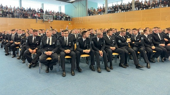 Polizeikräfte sitzen auf Stühlen in einem Saal.