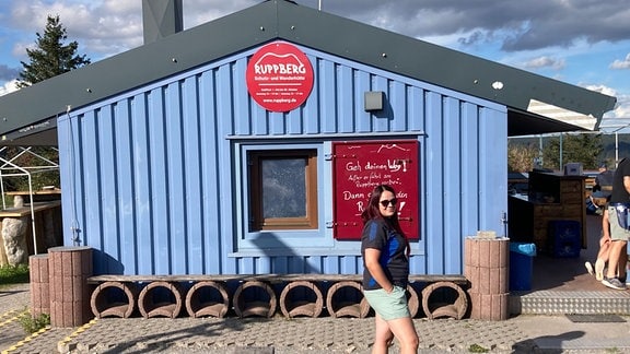 Steffi vom Ruppberg e.V. in Thüringen: Eine Frau mit kurzen Hosen, T-Shirt und Sonnenbrille steht vor einer blauen Hütte