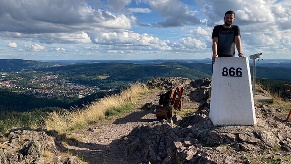 Philipp vom Ruppberg e.V. in Thüringen: Ein Mann steht auf einer Höhenmarkierung auf einem Berg über ihm blauer Himmel mit weißen Wolken, darunter weiter Blick über die Landschaft mit Hügeln und Wäldern