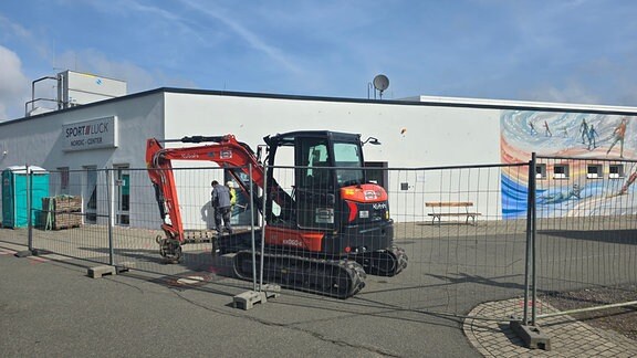 Ein Bagger und zwei Bauarbeiter befinden sich hinter einem Bauzaun.