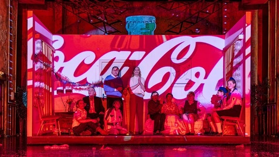 Auf der Bühne des Staatstheaters Meiningen: Darsteller werden von einer roten Coca-Cola-Werbung angestrahlt