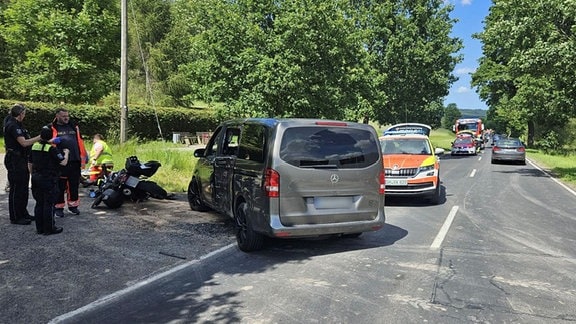 Helfernde stehen bei einem Unfall am Straßenrand, ein Kleinbus und im Hintergrund ein umgefallenes Motorrad.
