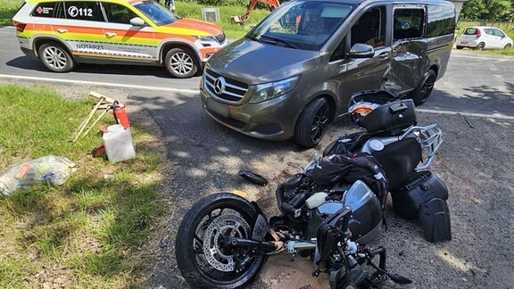 Ein kaputtes Motorrad und ein beschädigter Kleinbus am Straßenrand nach einem Unfall, im Hintergrund ein Notarztwagen.
