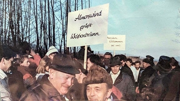 Altes Foto einer Menschmasse mit vielen fröhlichen Menschen in Winterklamotten und zwei Schildern "Almerswind grüßt Weißenbrunn" und "Herzlich willkommen bei uns"