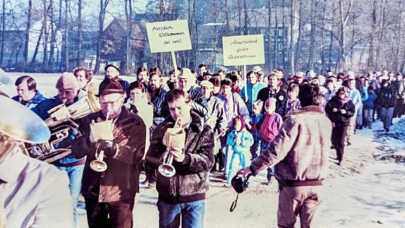 Altes Foto eines Stroms an Menschen in Winterklamotten und zwei Schildern "Almerswind grüßt Weißenbrunn" und "Herzlich willkommen bei uns", vorne läuft eine Blaskapelle voran