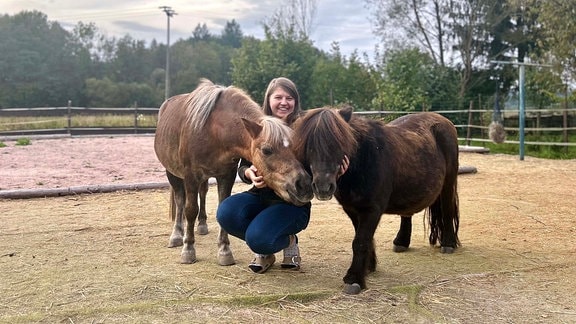 Eine lachende Frau kniet zwischen zweu Ponys auf einem ausgestreuten Platz 