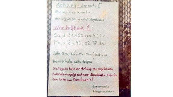 Altes Foto von handgeschriebenem Zettel mit Text: "Achtung - Einsatz! Endlich ist es so weit - der Signalzaun wird abgebaut! Wer hilft mit? Samstag, den 31.3.1990, ab 9 Uhr und Montag, 2.4.1990 ab 18 Uhr. Bitte 13er, 17er und 19er Schlüssel und Handschuhe mitbringen! Die Abgabe bzw. der Verkauf von abgebauten Materalien erfolgt erst nach Abschluß der Arbeiten. Ich bitte um Verständnis! Bauersachs - Bürgermeister -"  