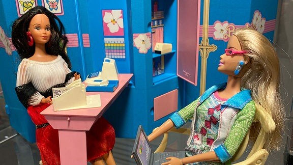 Barbie-Puppen in einer Ausstellung, eine sitzt auf einem Stuhl, die andere an einem Schreibtisch.