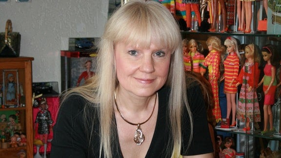 Eine Frau mit blonden Haaren ist vor einem Regal mit Barbie-Puppen zu sehen.