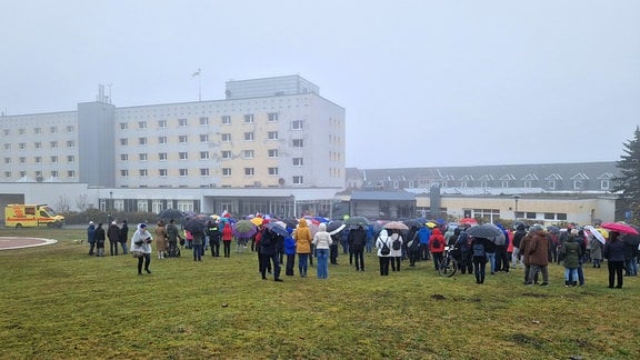 Menschen stehen im Nebel und Regen mit Schirmen vor dem Krankenhaus Neustadt.