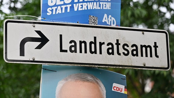 Plakate mit Fotos der Kandidaten hängen neben einem Wegweiser zum Landratsamt. 