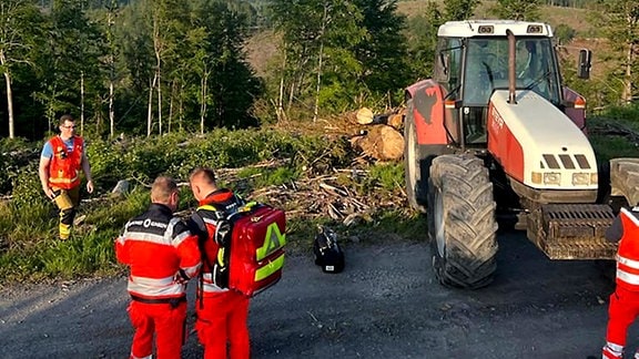 Rettungssanitäter stehen in einem Waldstück, dahinter ein Traktor und gefällte Bäume