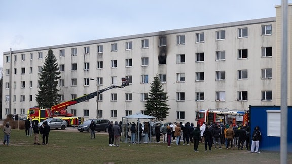 Menschern und Feuerwehrwagen mit Leiter vor einem mehrstöckigen Gebäude.
