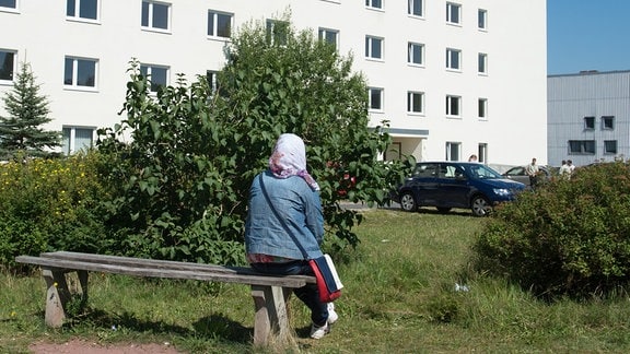 Ein Frau sitzt vor einem Wohnheim auf einer Bank.