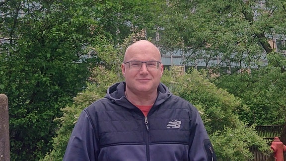 Ein Mann mit Brille und Sportjacke steht vor einem mehrstöckigen Gebäude.