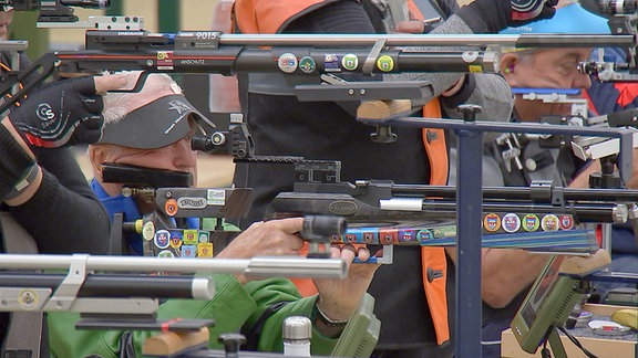 Ein Sportschütze zielt bei einem Wettbewerb mit einem Gewehr. Um ihn herum sind mehrere Gewehere.