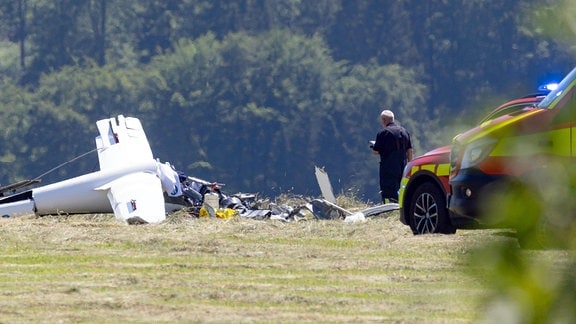 Ein Mann steht auf eienr Wiese vor einem Flugzeugwrack. Hinter ihm stehen zwei Feurwehrautos.