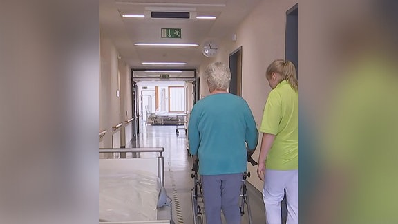 Eine Seniorin geht mit einer Pflegerin einen Gang entlang