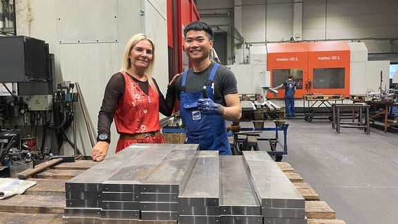 Katrin Fischer und Vin stehen in der Halle der Schmalkaldener Maschinenmesser GmbH. Vor ihnen auf einem Tisch liegen gestapelte Metallteile.