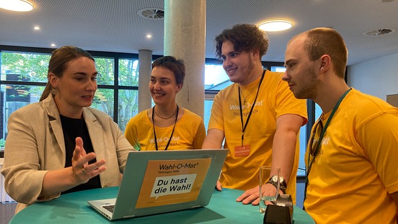 Drei Menschen in gelben T-Shirts stehen an einem Laptop an einem Tisch