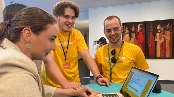 Drei Menschen in gelben T-Shirts stehen an einem Laptop an einem Tisch