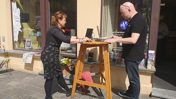 Eine Frau und ein Mann stehen draußen vor einem Ladenlokal an einem Tisch mit ihren Laptops. Sie schauen auf die Bildschirme der Laptops.