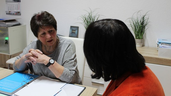 Eine Frau im grauen Pullover und eine Frau im orangen Jackett sitzen an einem Tisch und unterhalten sich