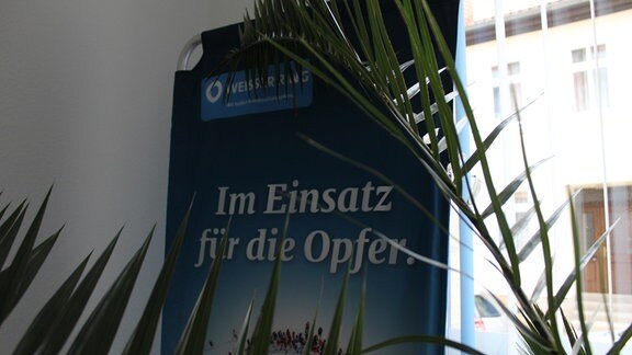 ein Transparent mit der Aufschrift "Im Einsatz für die Opfer" steht hinter einer Palme