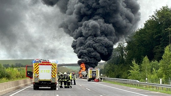 Im Vordergrund ist ein Löschfahrzeug mit Einsatzkräften. Im Hintergrund ein Brennender Tanklastwagen auf der Autobahn.