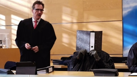 Ein Angeklagter vor Gericht verbirgt sein Gesicht hinter einem Aktenordner.