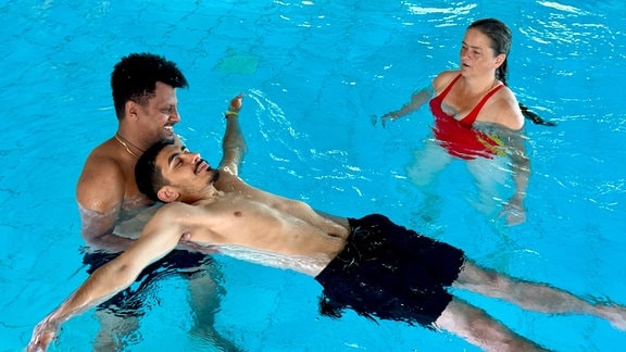 Drei Menschen in einem Schwimmbecken.Ein Mann mit schwarzer Badehose liegt mit ausgestreckten Armen auf der Wasseroberfläche und wird von einem anderen Mann gehalten, damit er nicht untergeht.